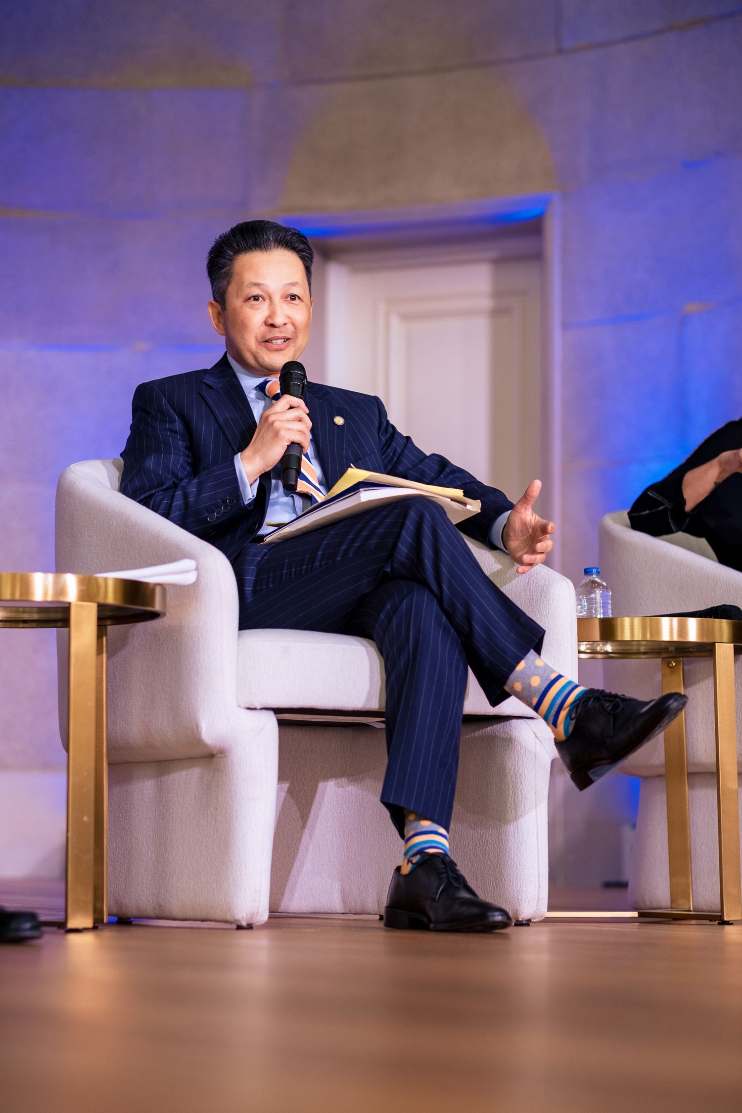 A man in a navy pinstripe suit with colorful socks speaks into a microphone while seated in a white chair on stage.