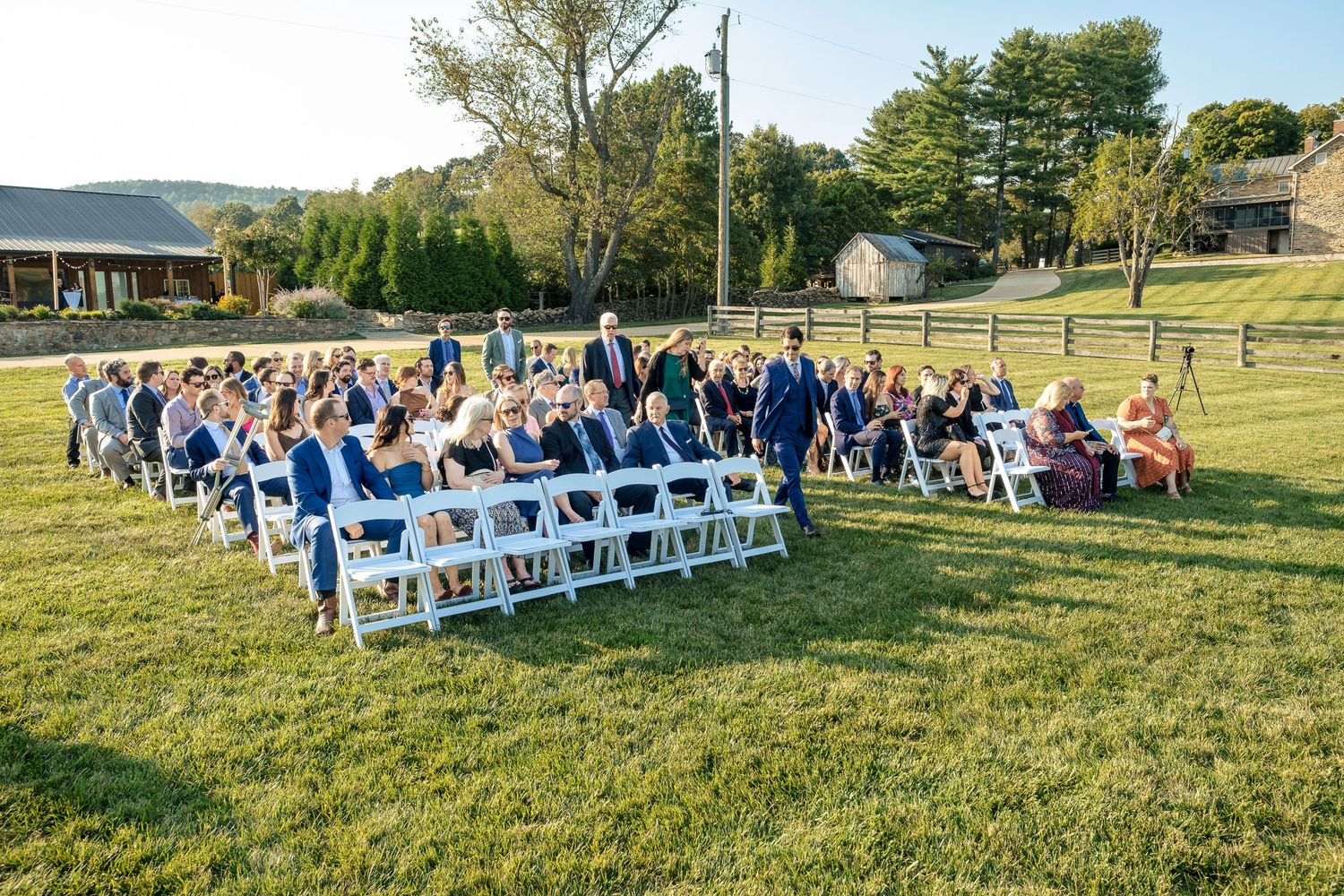 Guests seated in white chairs on a green lawn for an outdoor wedding ceremony in a scenic countryside setting.