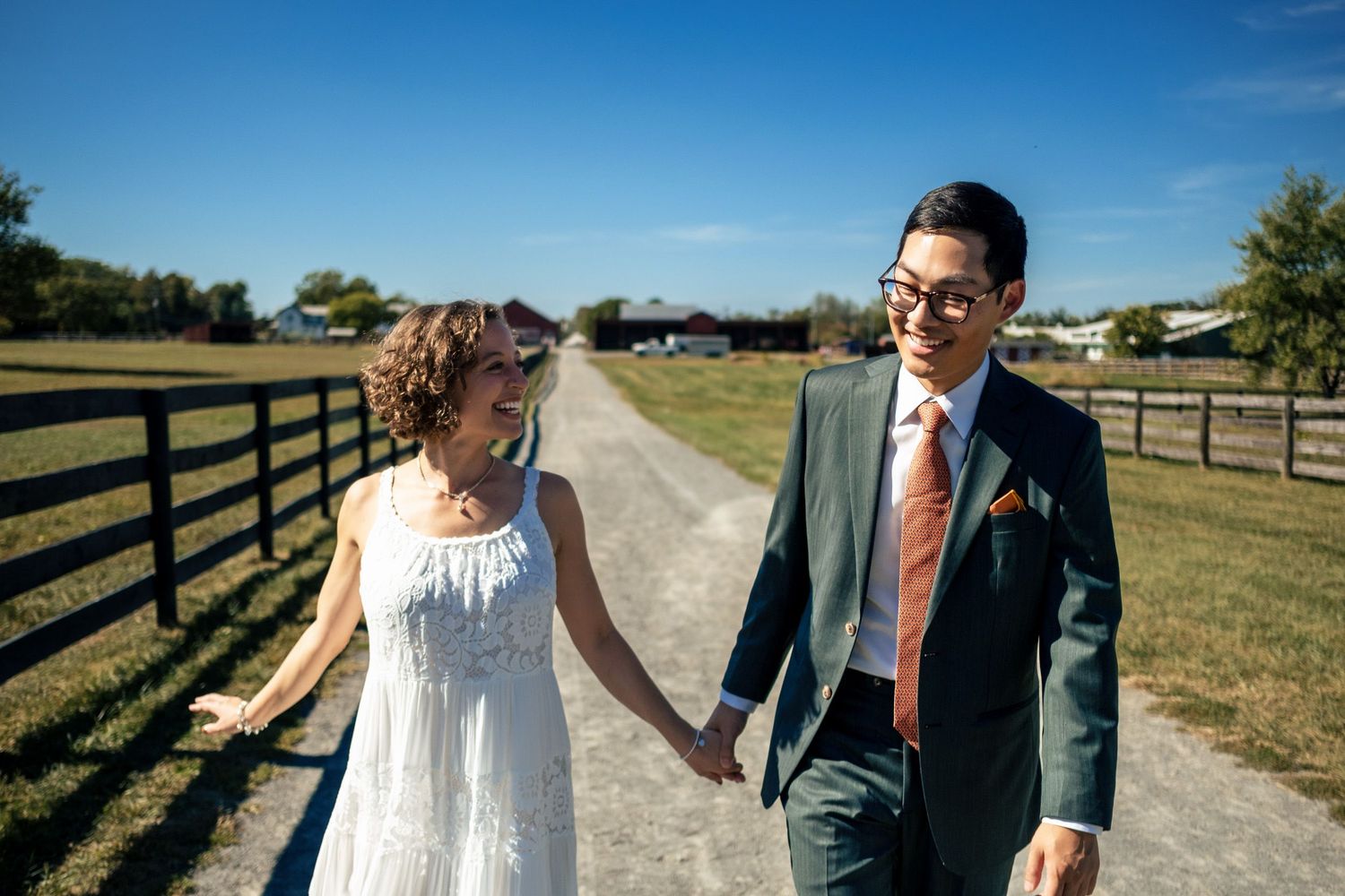Happy newlyweds holding hands and walking down a sunny farm path lined with wooden fencing.
