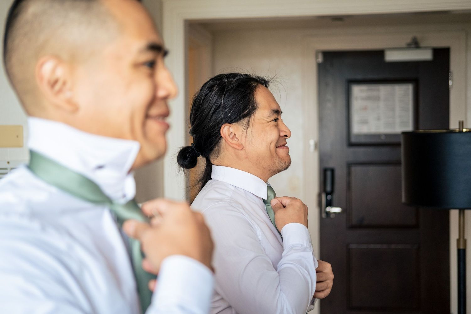 Two men in white dress shirts smiling while adjusting green ties during wedding preparations.
