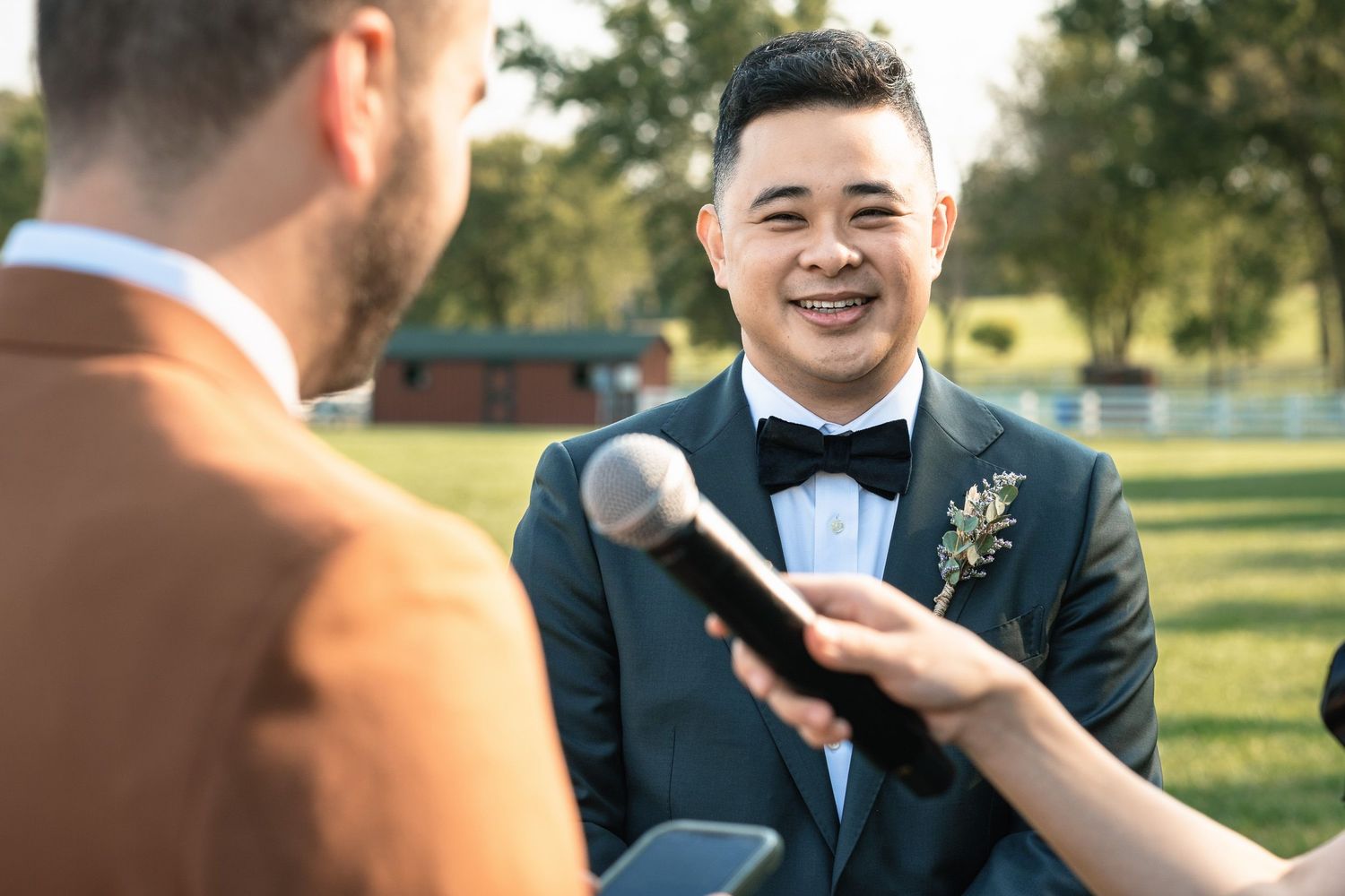 Groom in dark tuxedo with boutonniere smiling joyfully during outdoor wedding ceremony on a farm.