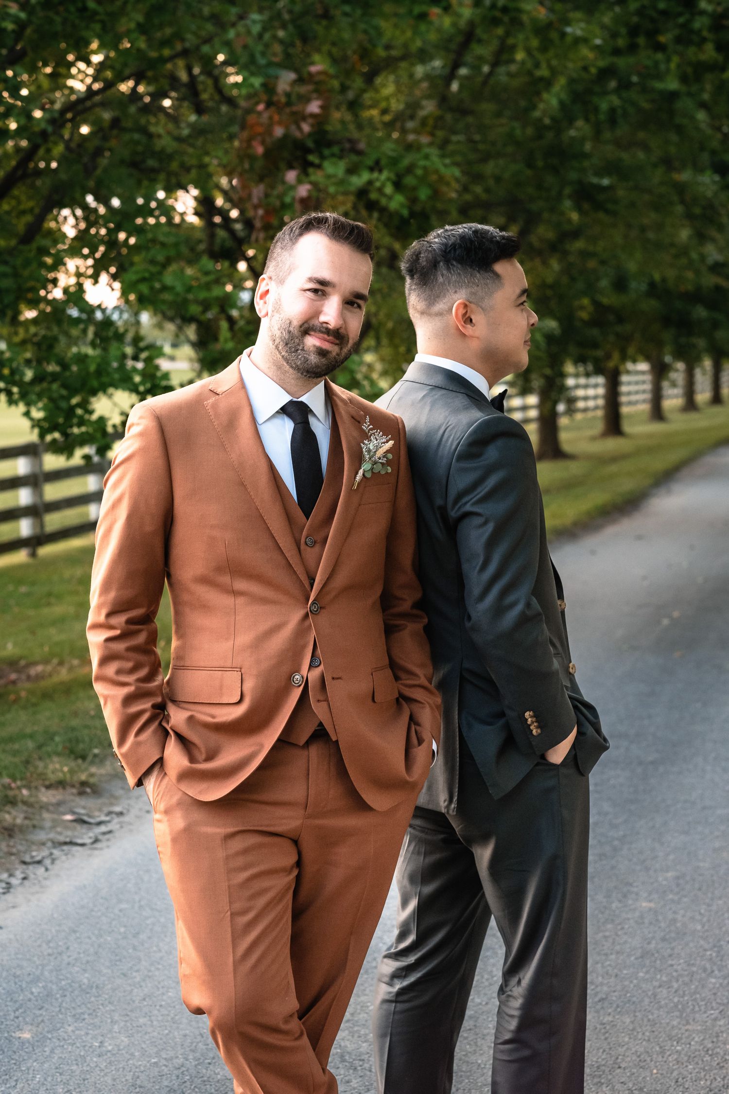 Groom in rust brown three-piece suit with boutonniere posing on tree-lined driveway at wedding.