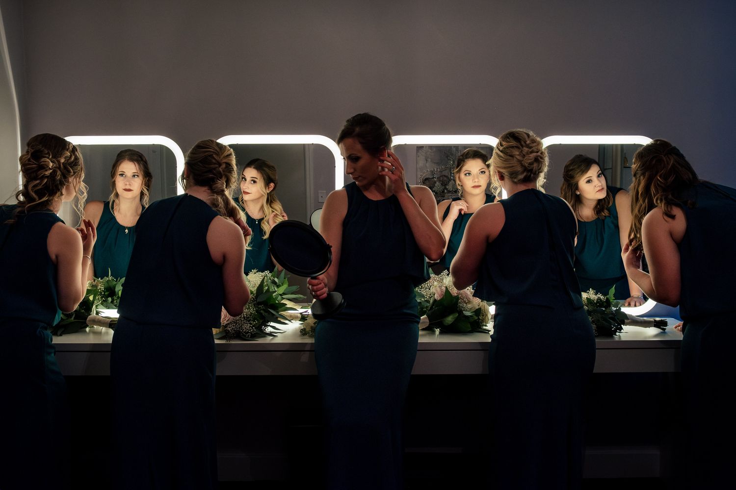 Bridesmaids in navy dresses apply makeup together at a lit vanity mirror.