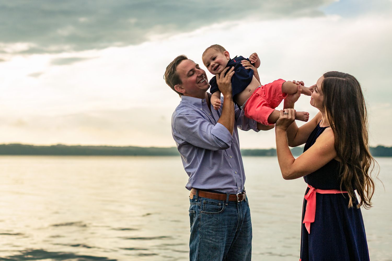 Happy family with baby enjoying sunset by the lake, parents lifting child together outdoors.
