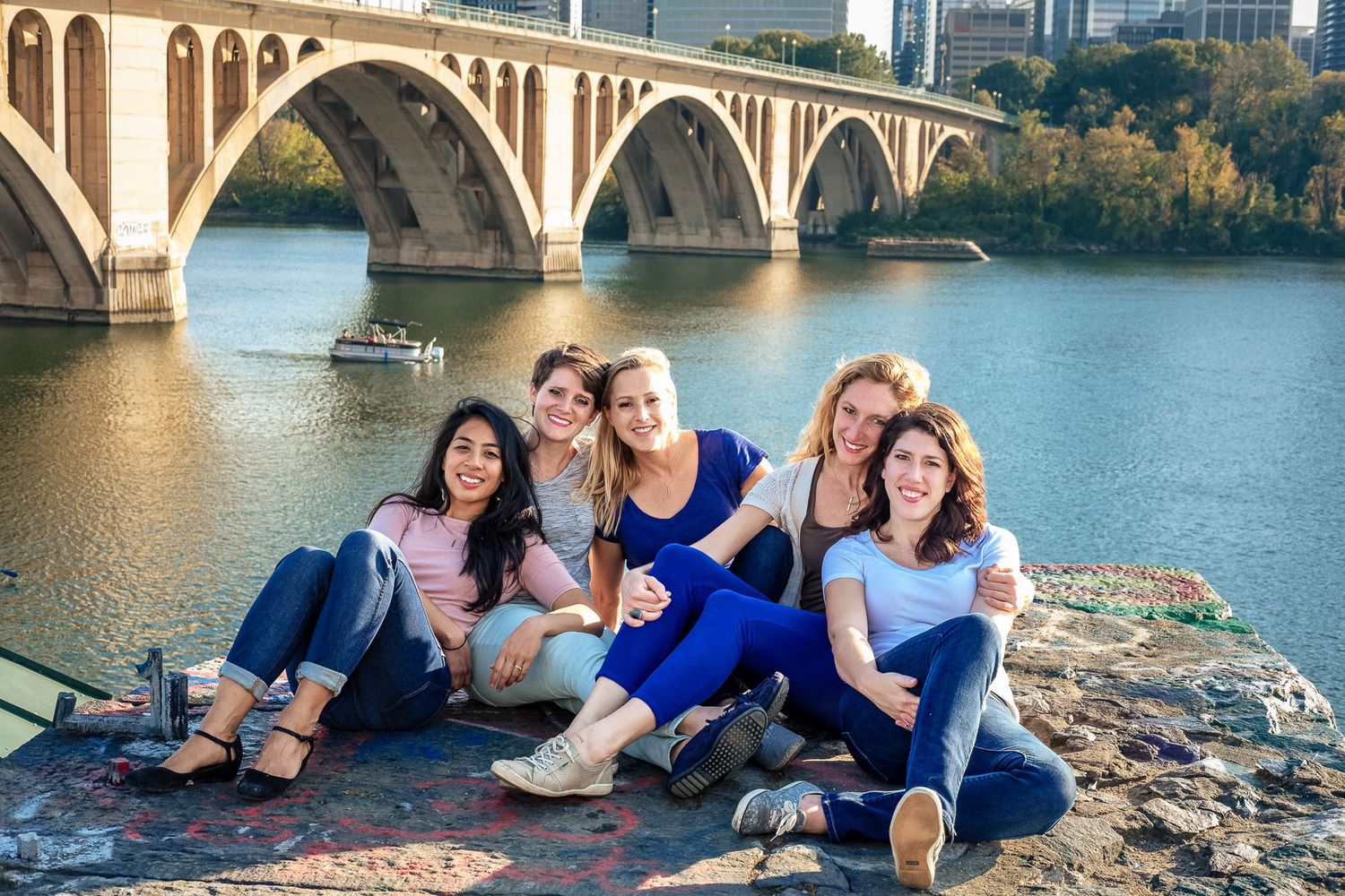 Group of five smiling women sitting together outdoors by a river with a bridge backdrop.