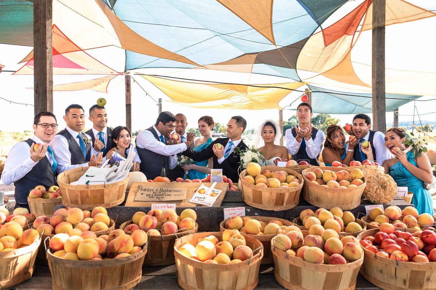 Wedding party posing joyfully behind colorful baskets of fresh peaches at an outdoor market.