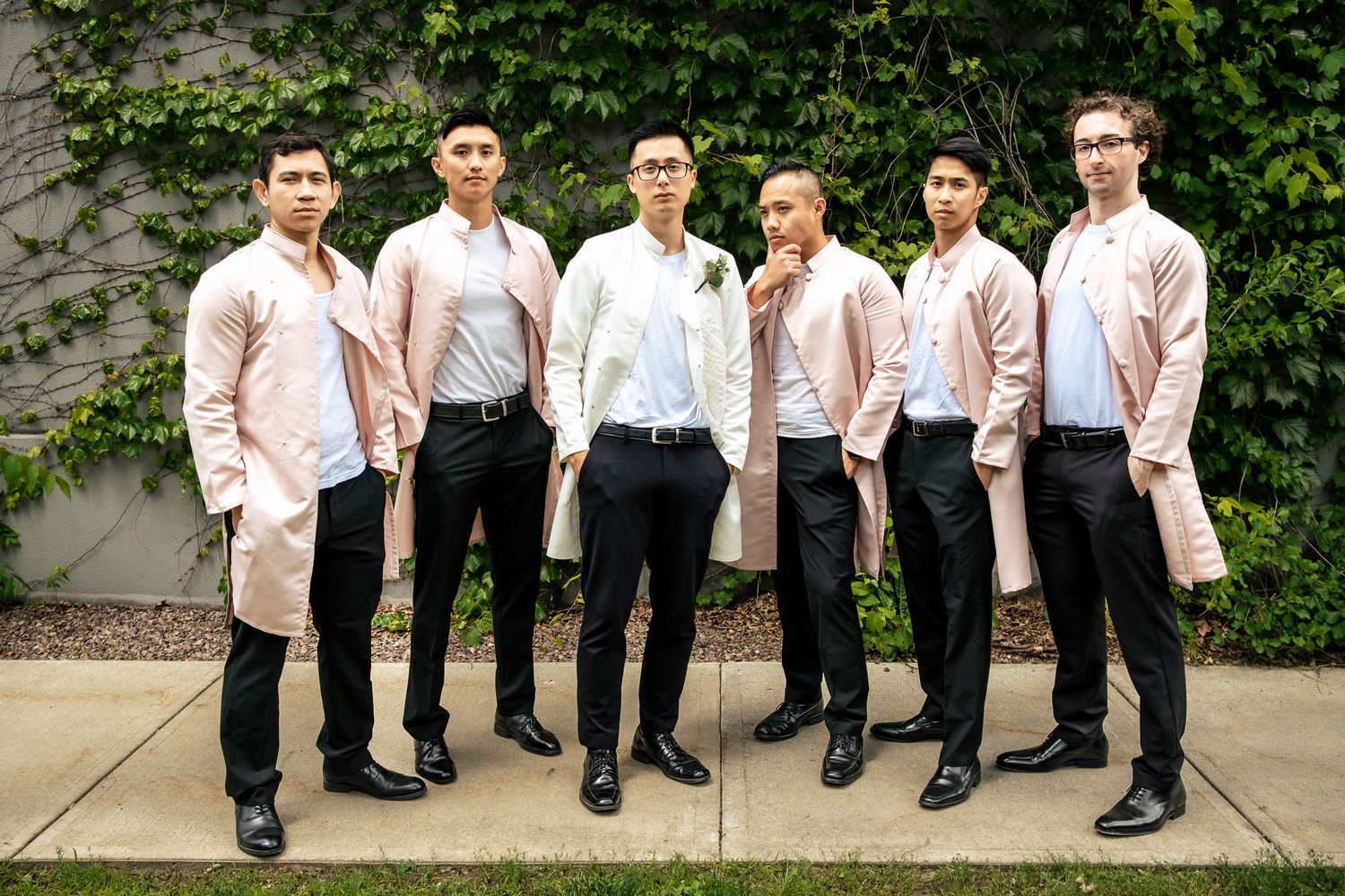 Groom and groomsmen posing in matching blush pink coats and black pants outdoors.