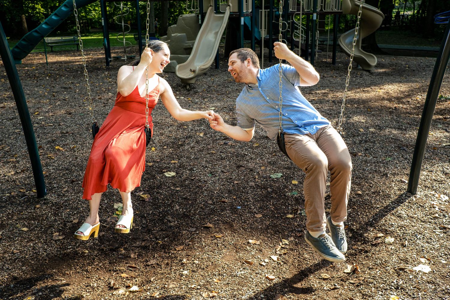 Playful couple holding hands on swings at a sunny playground, woman in red dress laughing.