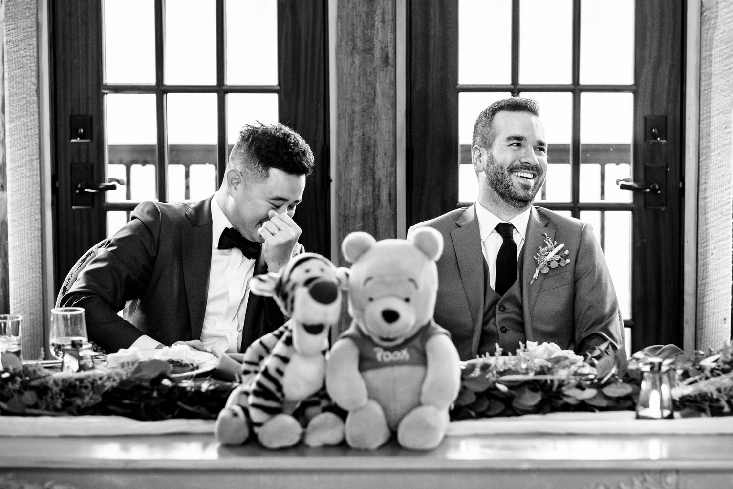 Two grooms sharing an emotional moment at wedding table with Pooh and Tigger plush toys.