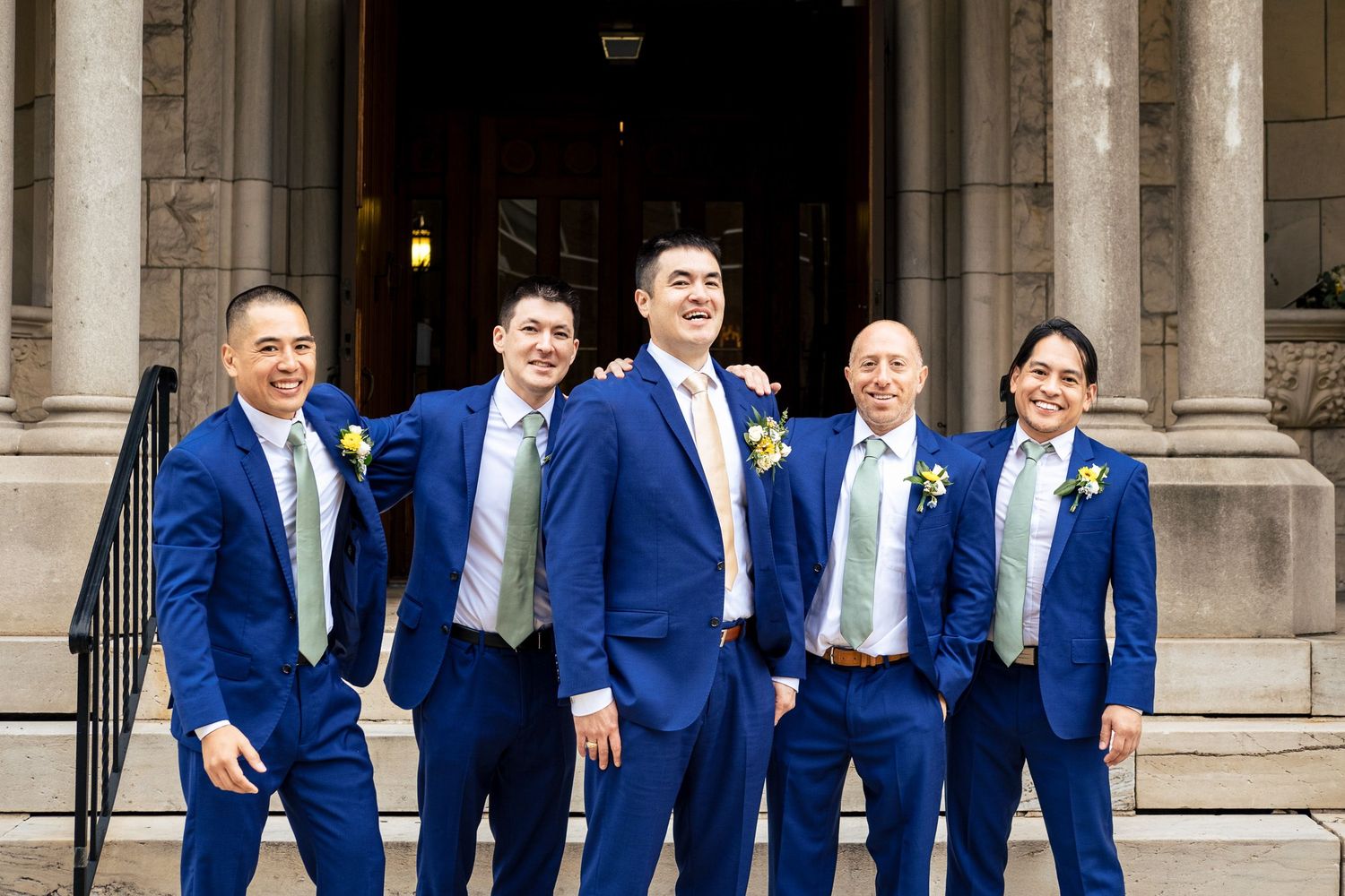 Five groomsmen in matching royal blue suits and sage green ties posing confidently on stone church steps.
