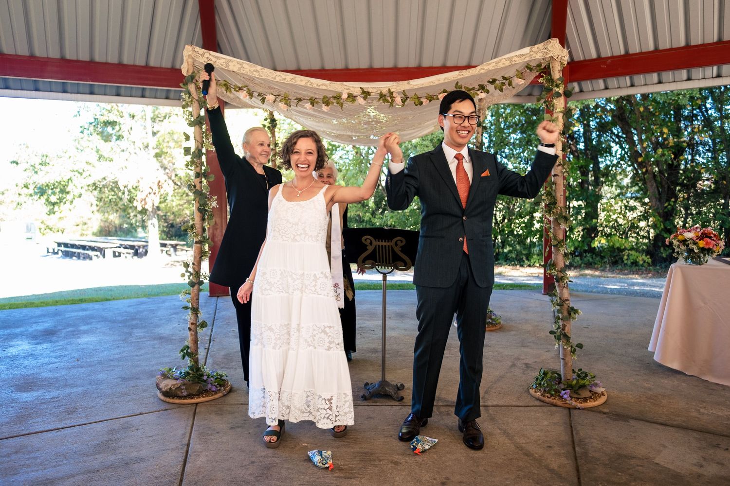 Bride and groom raise hands in celebration with officiant under decorated wedding chuppah outdoors.