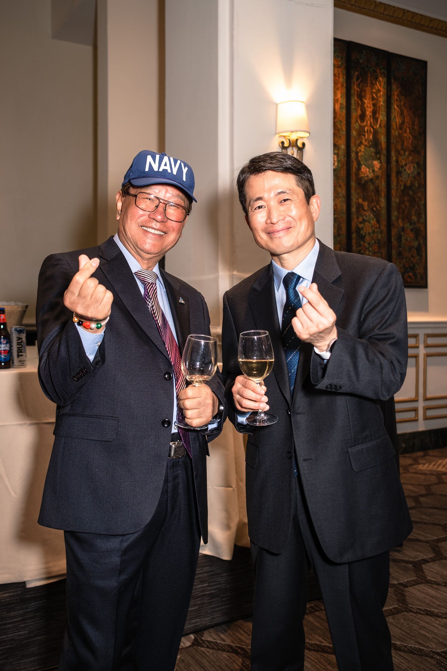 Two men in suits holding wine glasses and making finger heart gestures, one wearing a Navy cap, at a formal event.