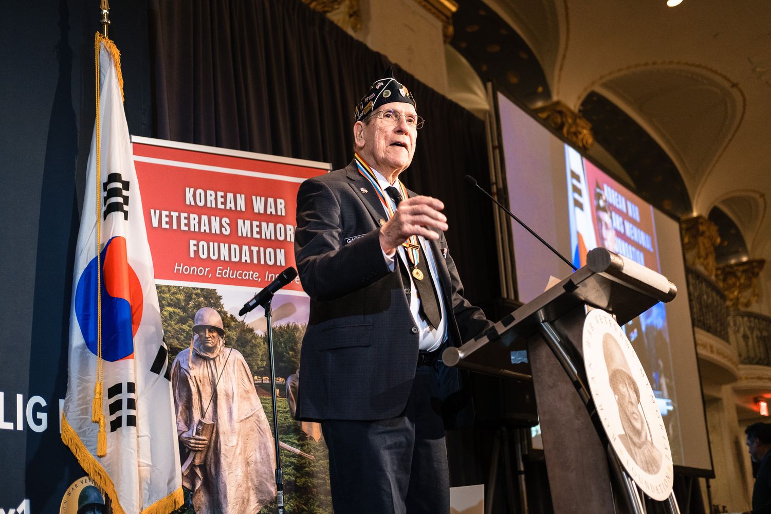 Speaker at Korean War Veterans Memorial Foundation event with South Korean flag and memorial backdrop.
