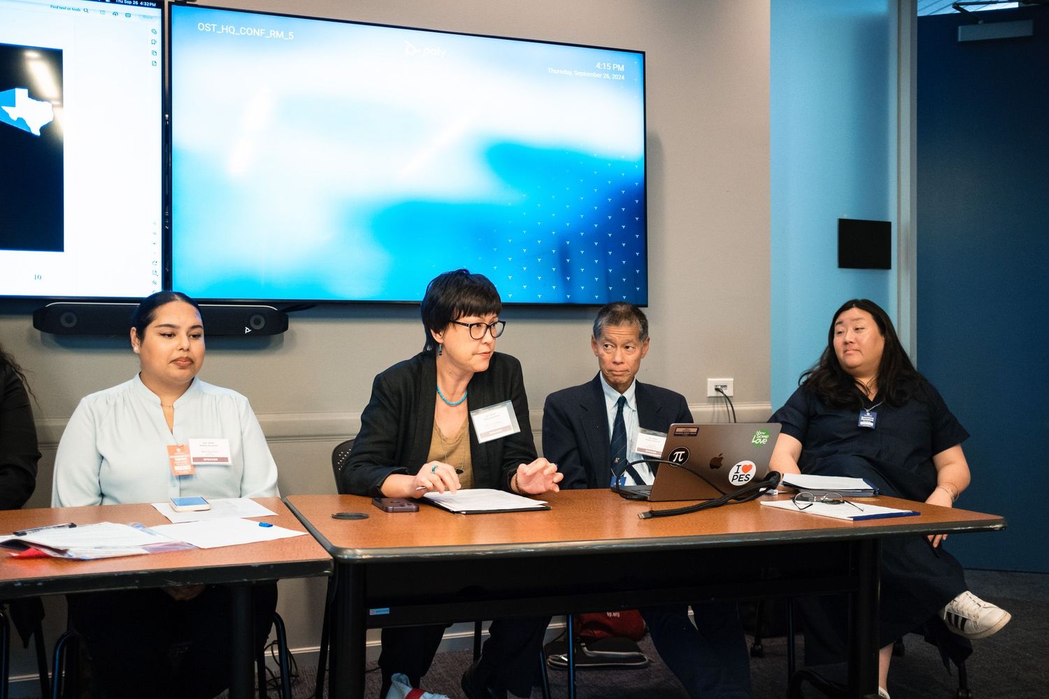 Four professionals seated at a conference table during a panel discussion with a large screen behind them.
