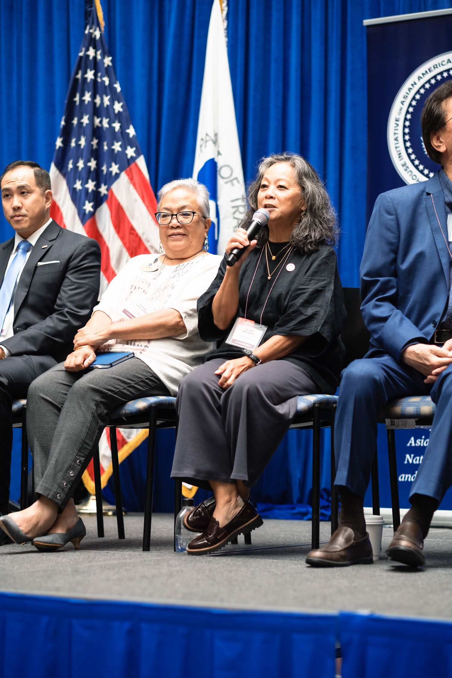 Panel of diverse speakers at official government event with US flag, woman speaking into microphone at formal conference.