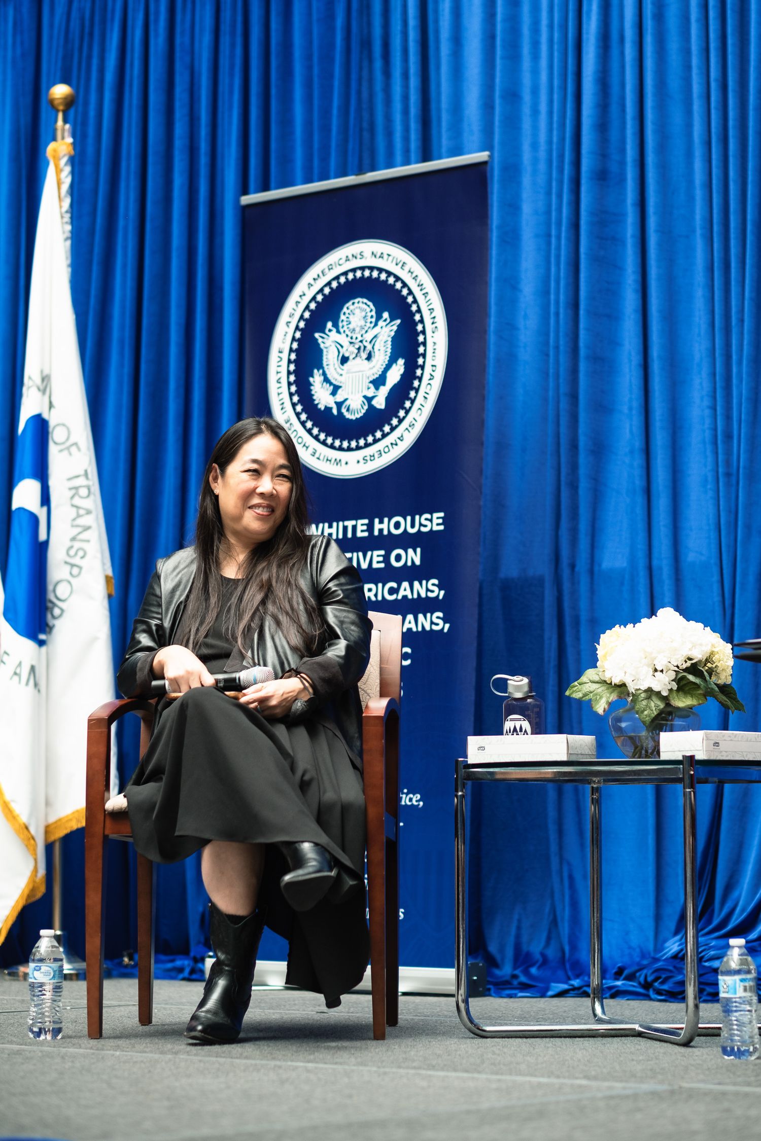 Woman seated on stage at White House initiative event, blue curtain backdrop with official seal banner.