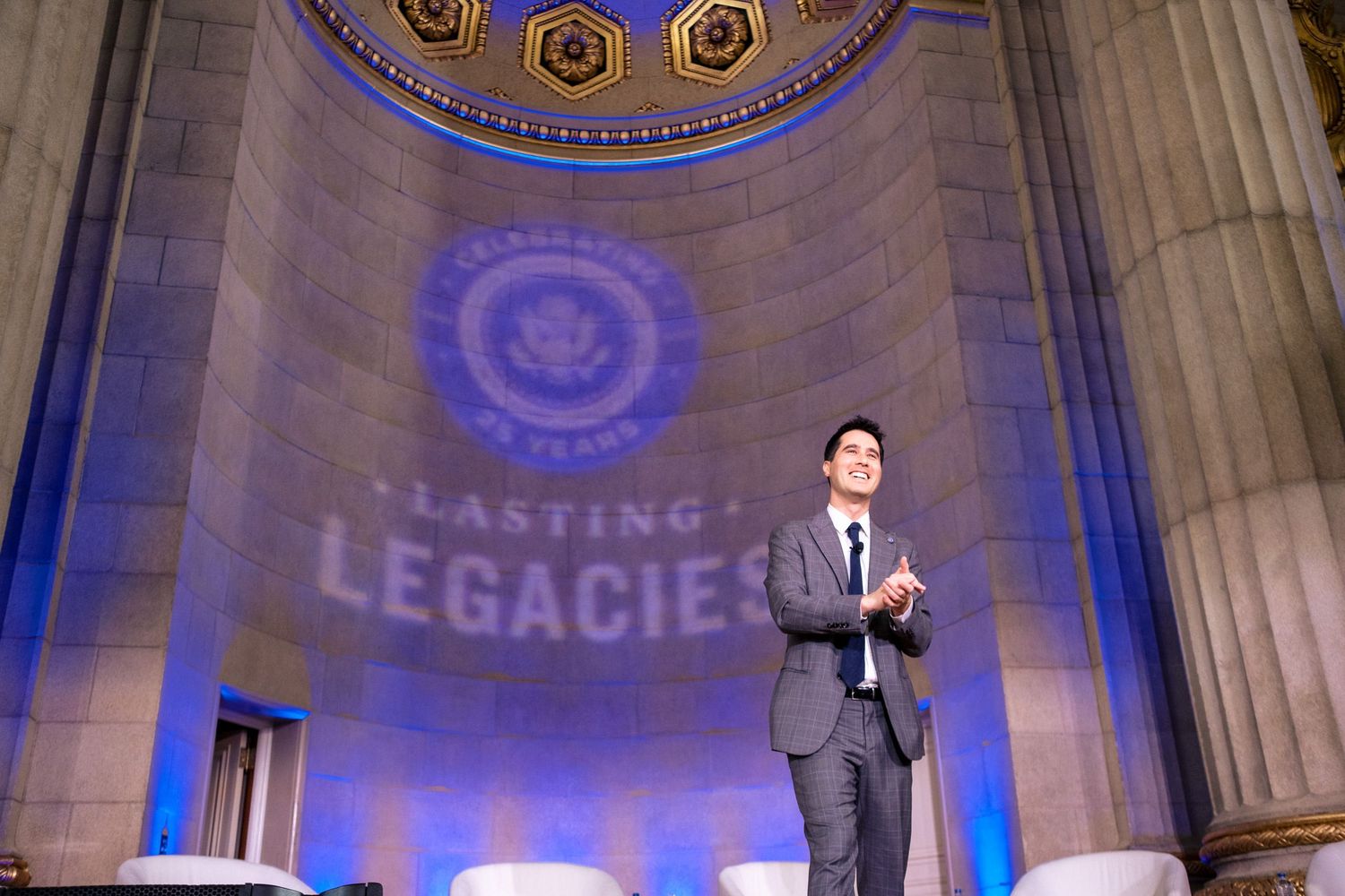 Man in suit stands on stage at Lasting Legacies event in grand rotunda with blue lighting and projected logo.