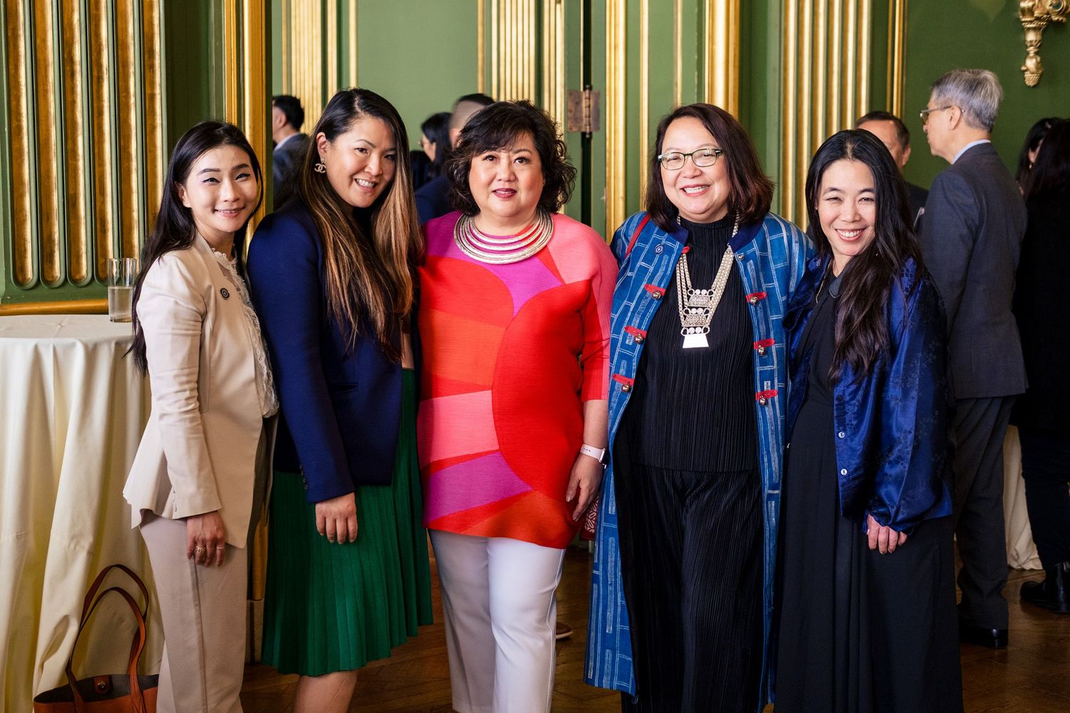Five women pose together smiling in an elegant green-walled ballroom at a formal event.