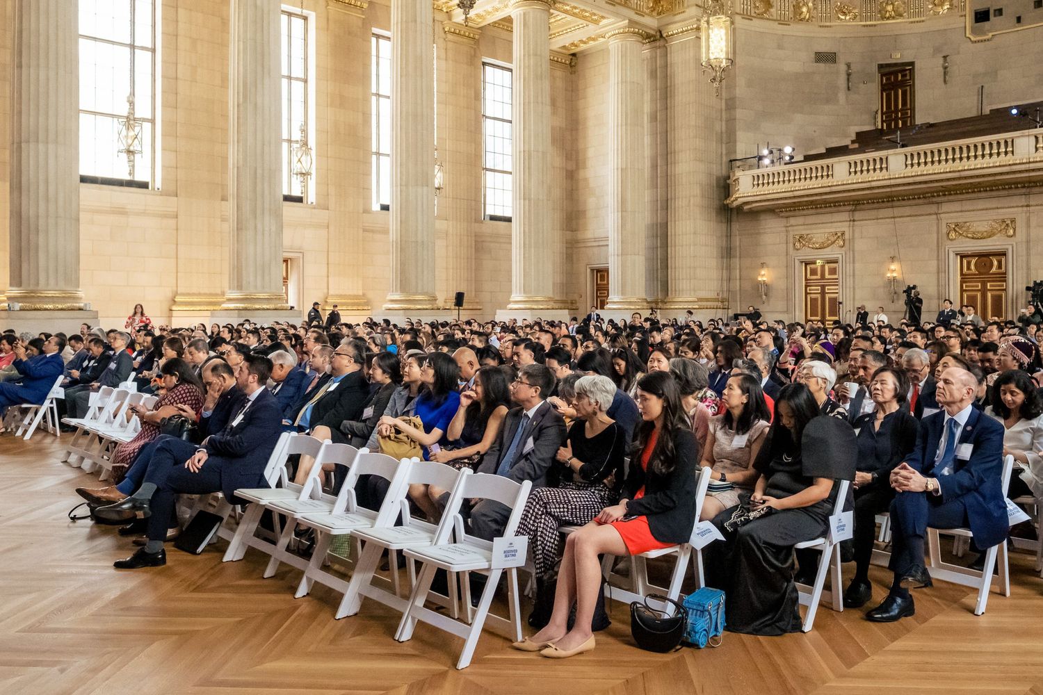 Large crowd seated in white chairs at a formal ceremony inside an ornate neoclassical hall.