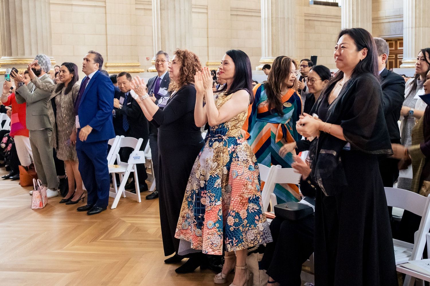 Diverse crowd standing and applauding at an indoor ceremony in a grand hall, many holding papers and phones.