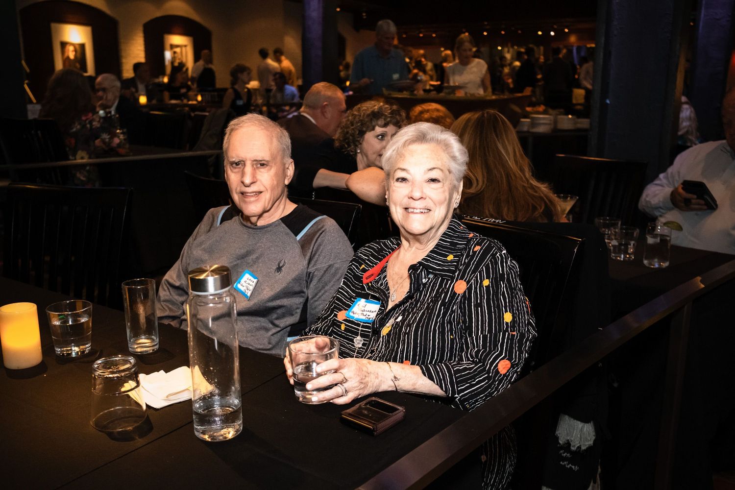 Two seniors smile at a bar table with drinks in a dimly lit venue.