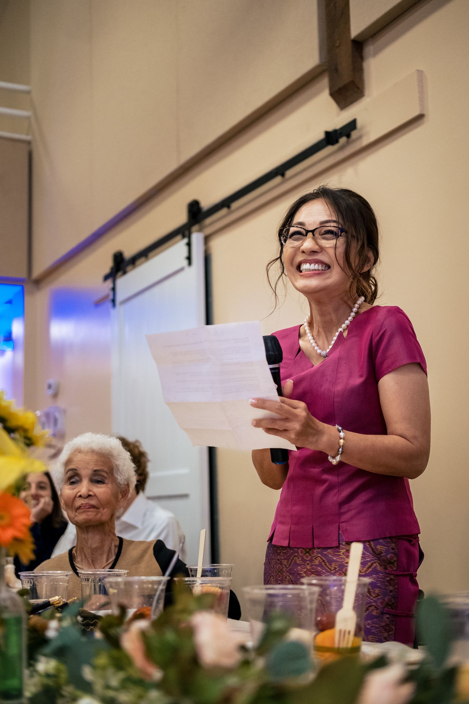 Woman in pink top giving a heartfelt speech at a wedding reception holding papers.