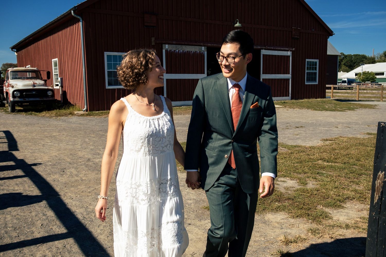 A happy newlywed couple walks hand-in-hand past a red barn on their wedding day.