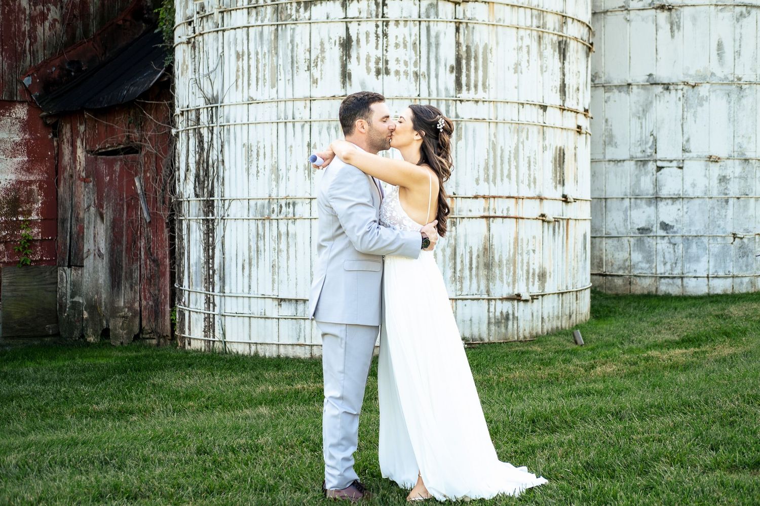 Bride and groom share a romantic kiss in front of rustic white grain silos at farm wedding.
