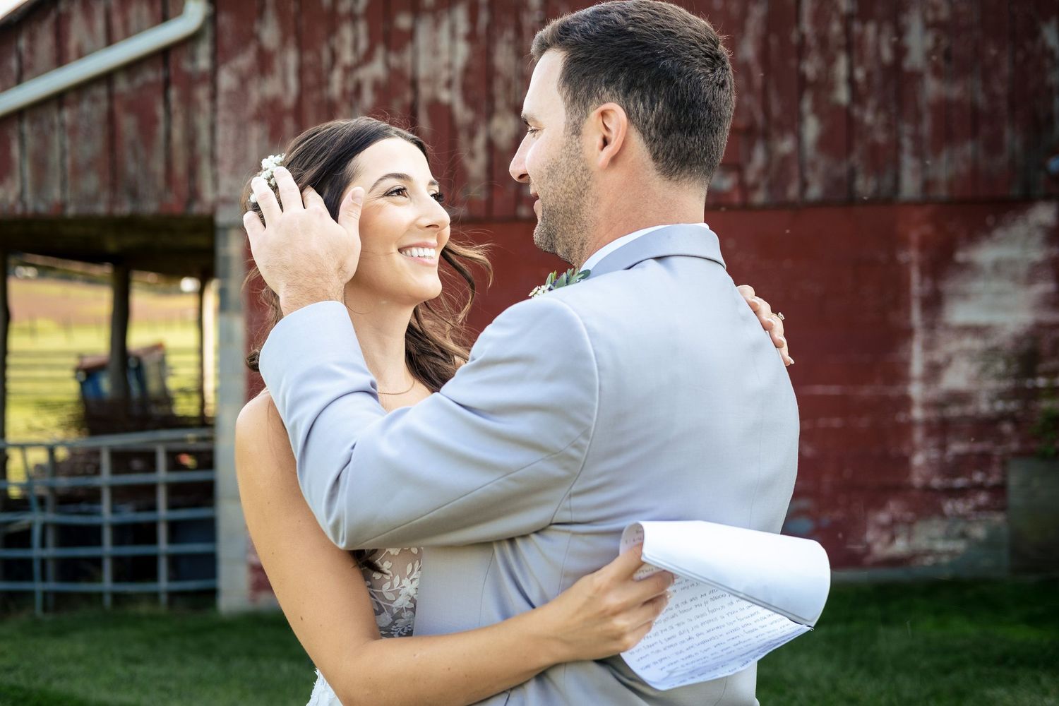 Smiling bride and groom embrace near a red barn during their farm wedding celebration.