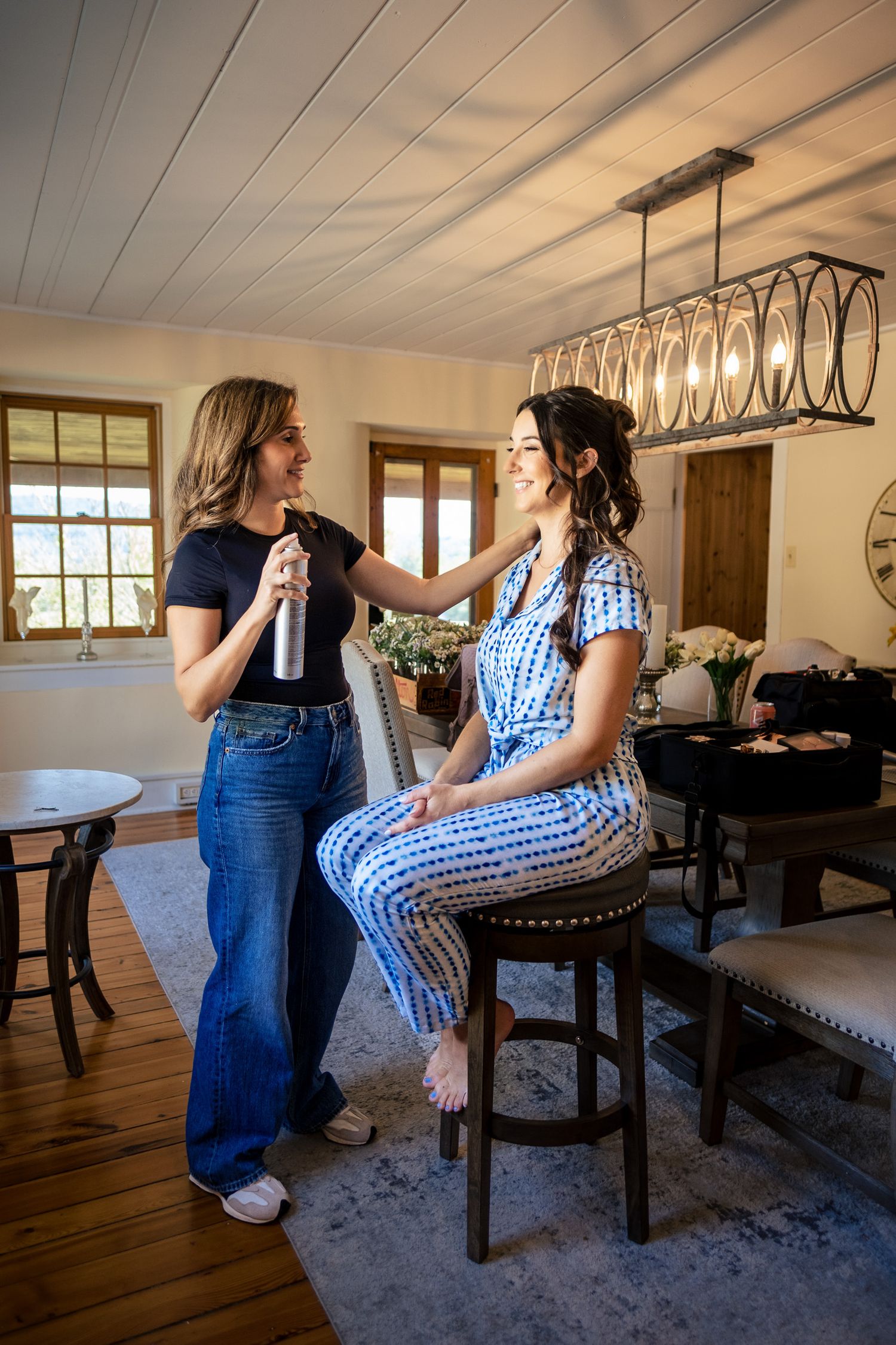 Hairstylist sprays bride's hair while she sits on a stool in a bright, elegantly decorated room.