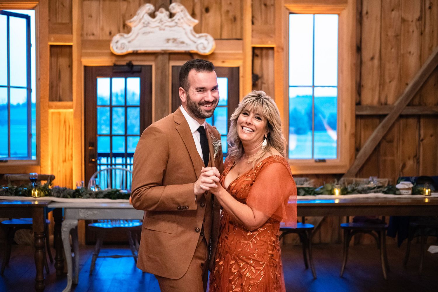 Groom in brown suit and woman in orange dress smile while holding microphone at barn reception.