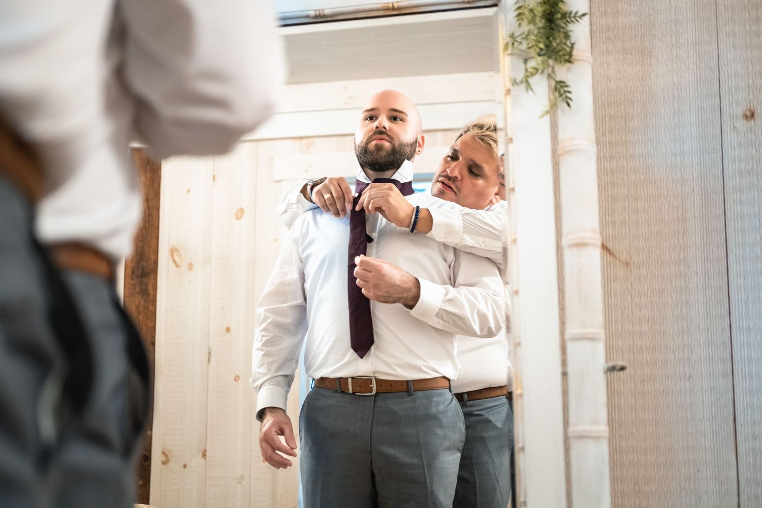 Woman helping bearded groom tie his necktie while standing before a mirror.