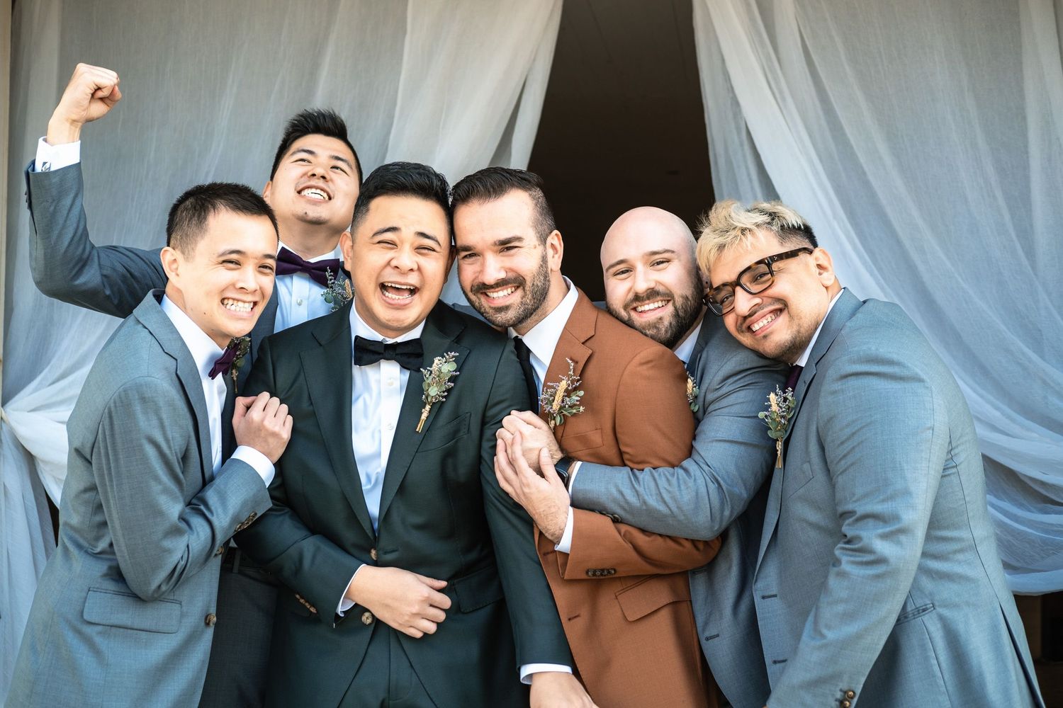 Two grooms celebrate with their groomsmen in matching gray suits at a barn wedding.