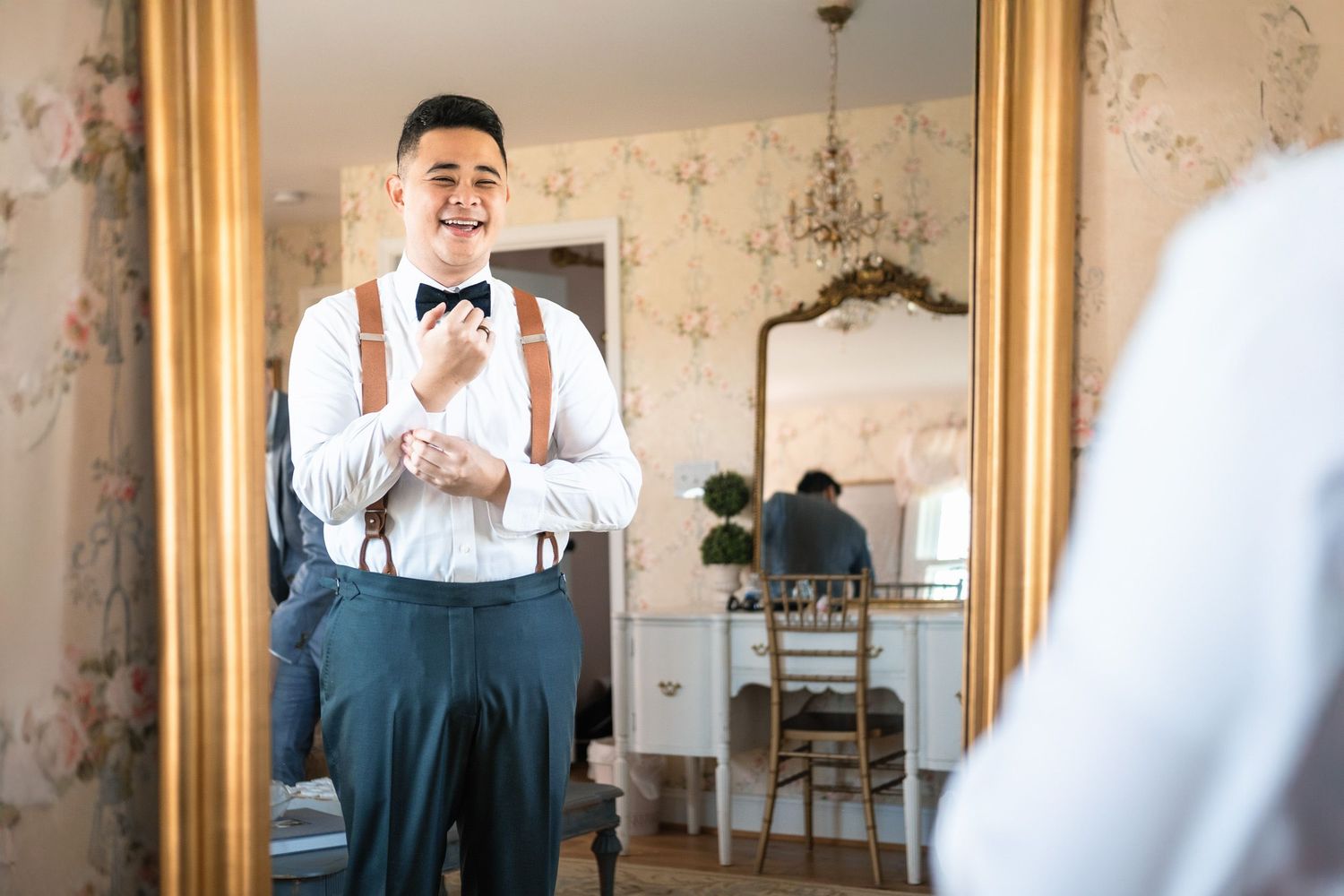 Groom smiling while adjusting bow tie in ornate gold mirror before wedding.