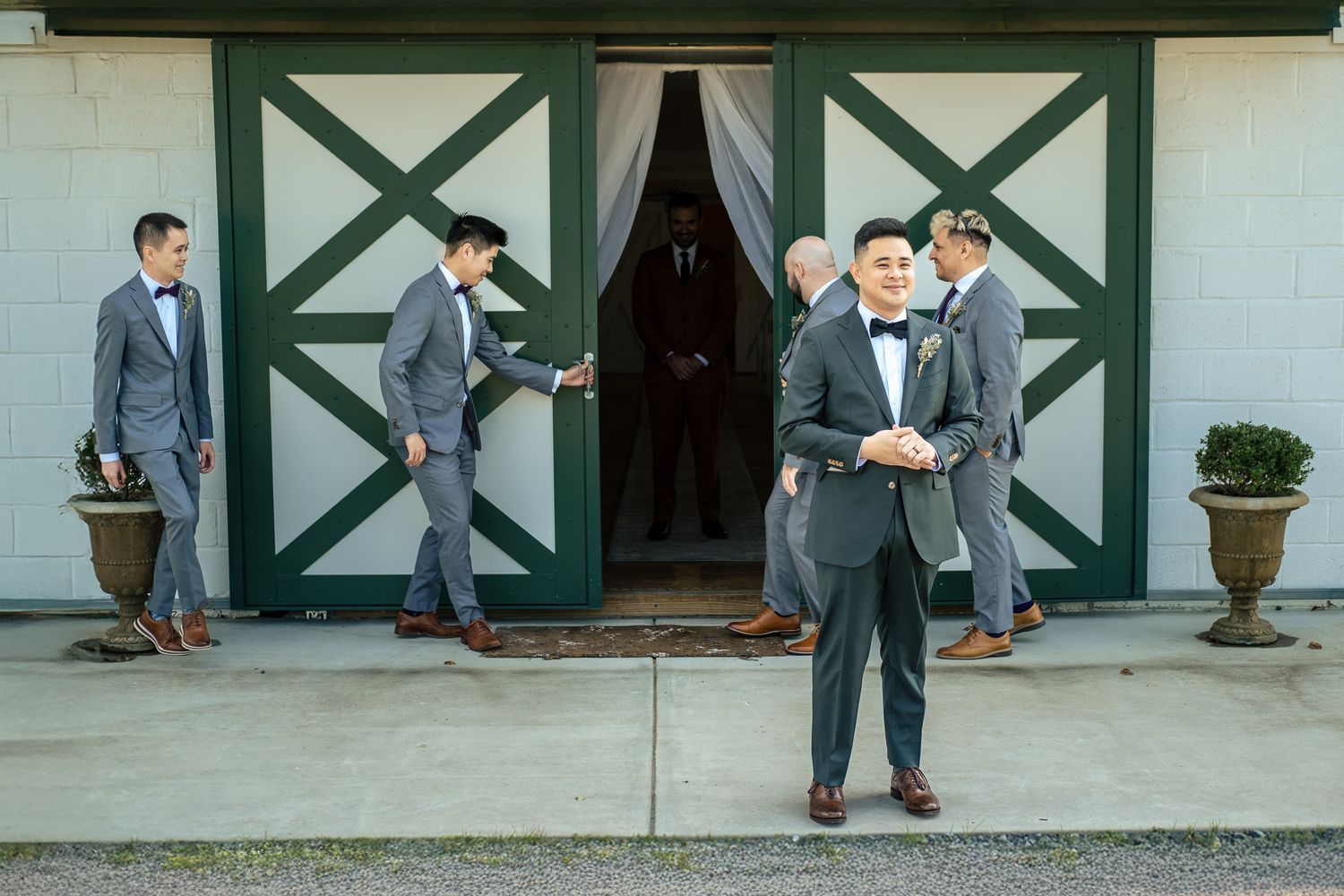 Groomsmen in gray suits and bow ties pose outside a white barn with green doors at a wedding.