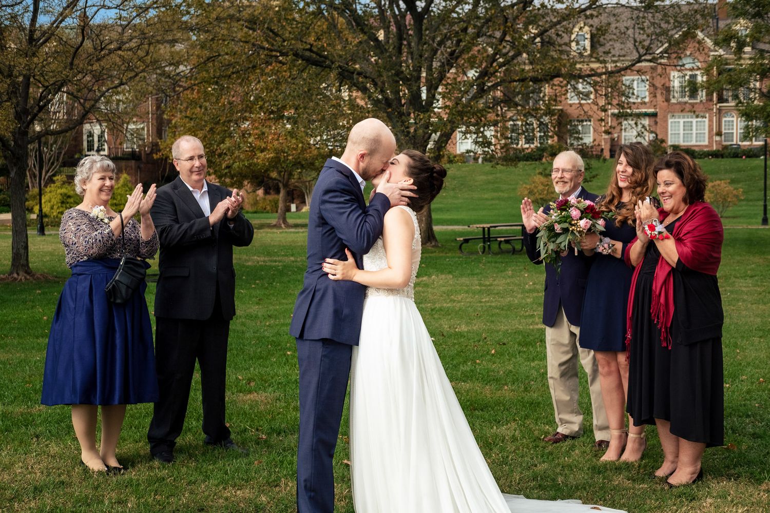 Newlyweds share their first kiss outdoors on a green lawn surrounded by cheering family members.