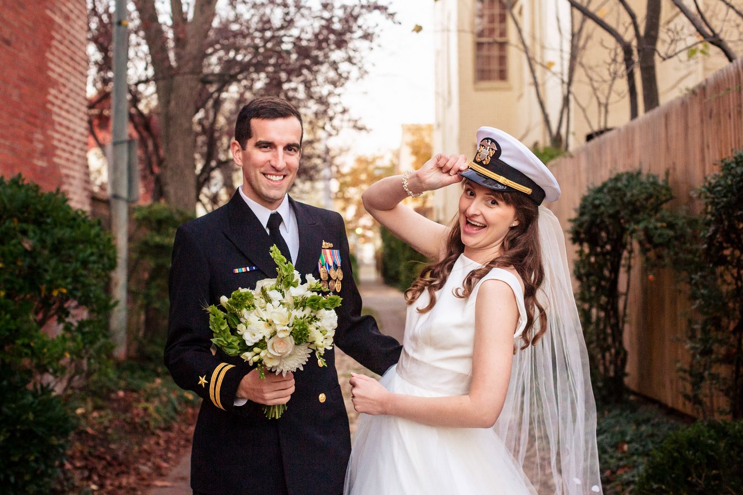 Naval officer groom and bride playfully pose with his military cap and white bouquet outdoors.