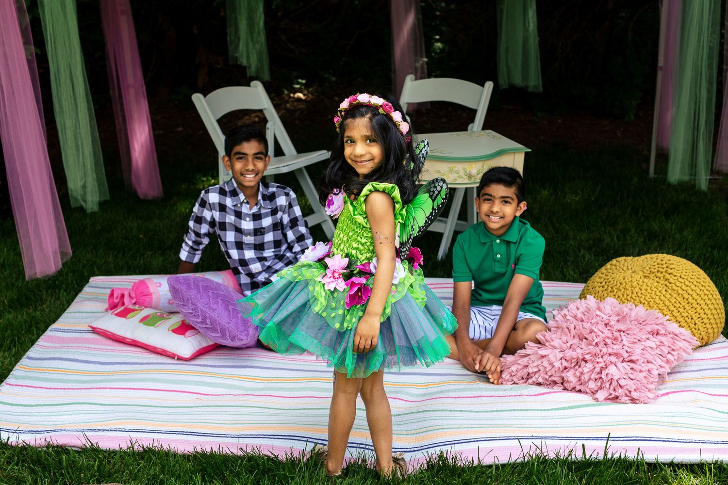Three children posing outdoors at a party, with a girl in a colorful green tutu dress flanked by two boys.