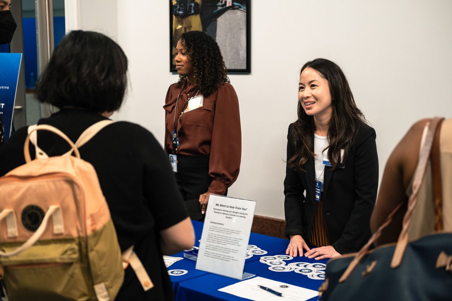 Two young women smile at a career fair table with a blue tablecloth, engaging with a visitor in a professional setting.