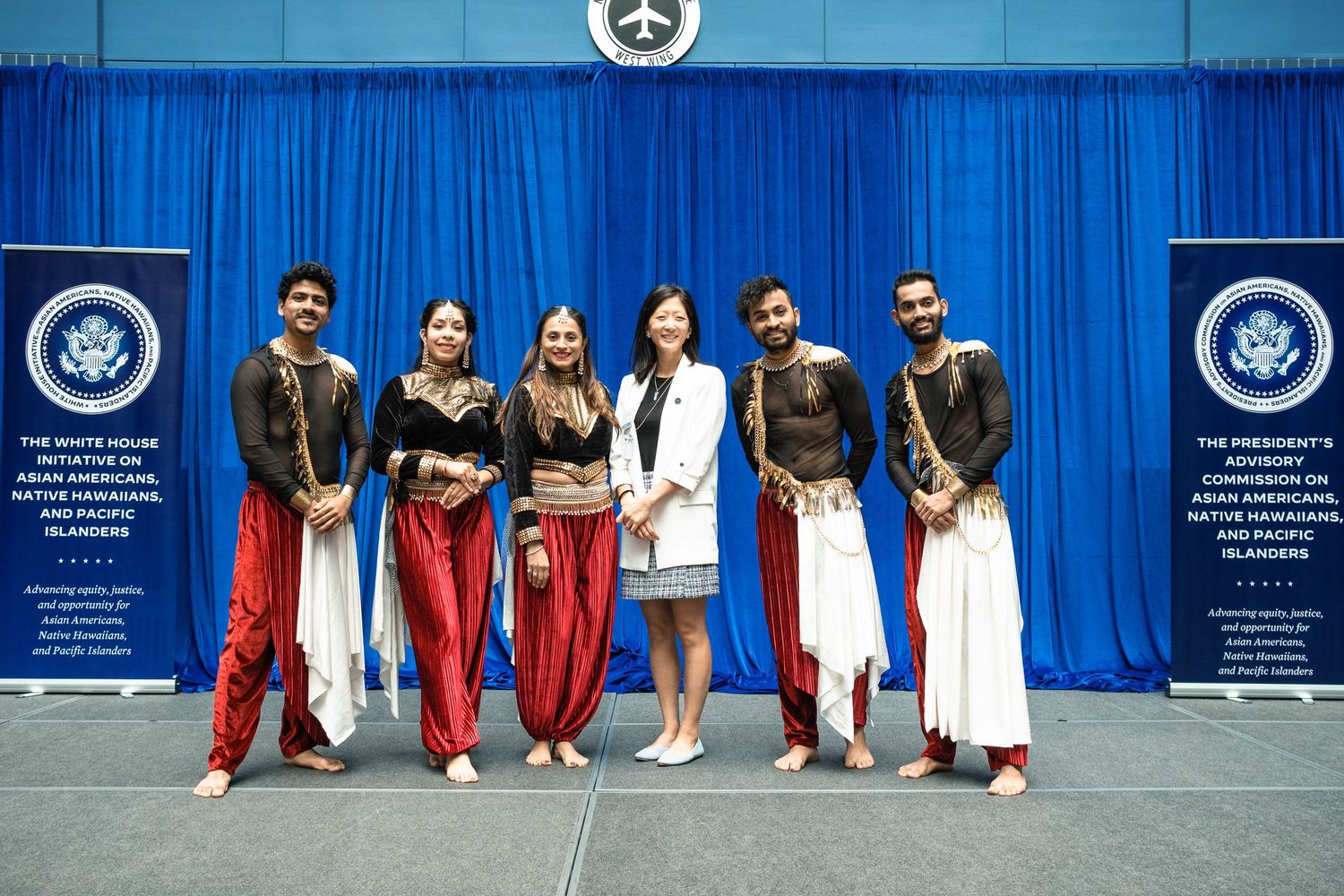 Five performers in traditional Indian dance costumes pose with woman at White House AANHPI initiative event.
