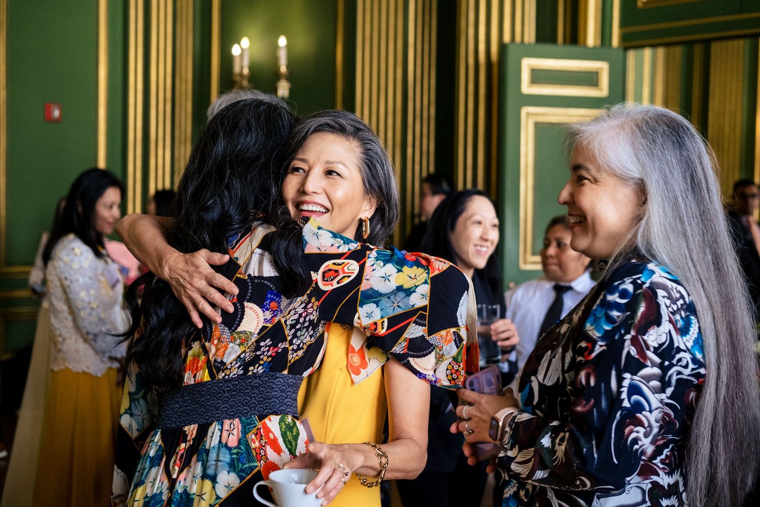 Women in colorful traditional attire embrace and socialize at an elegant green-walled formal reception event.