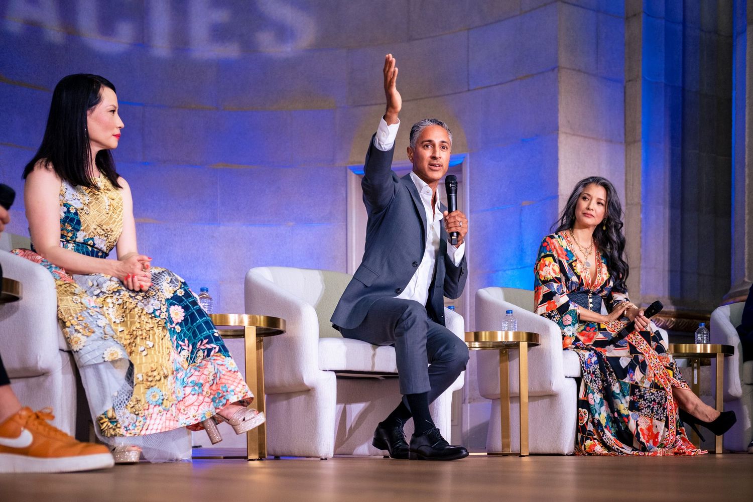 Three panelists on stage, man raising hand with microphone, two women in floral dresses seated beside him.