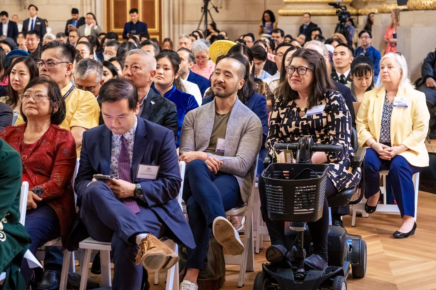 Diverse audience seated at a formal indoor event, including a woman in a wheelchair, in an elegant hall.