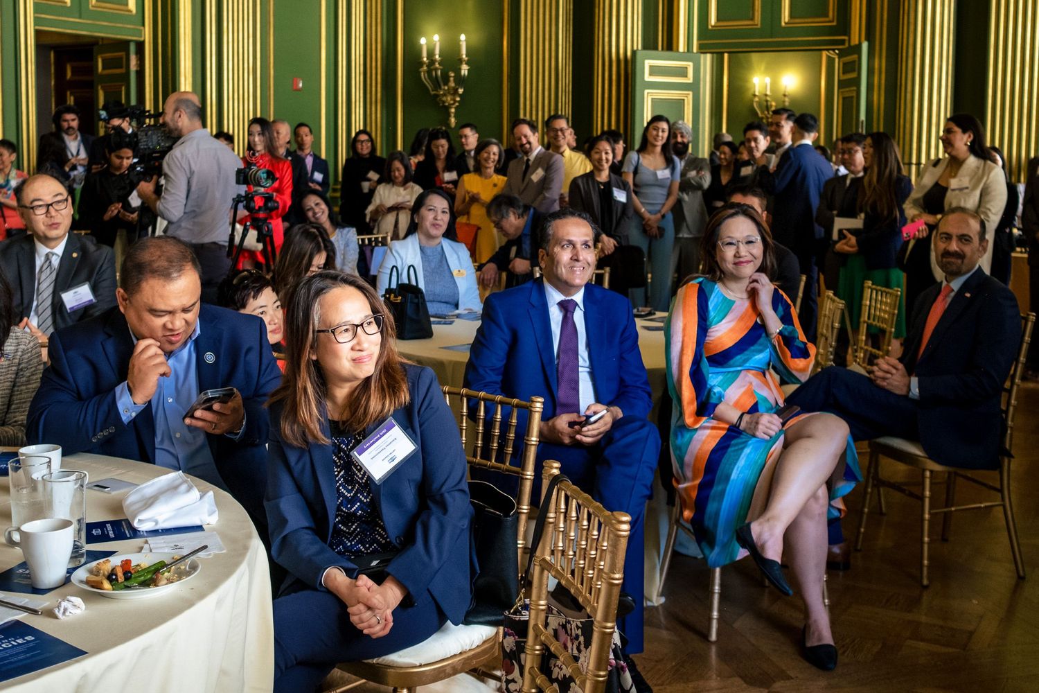 Attendees seated at round tables during a formal conference event in an elegant green and gold ballroom.