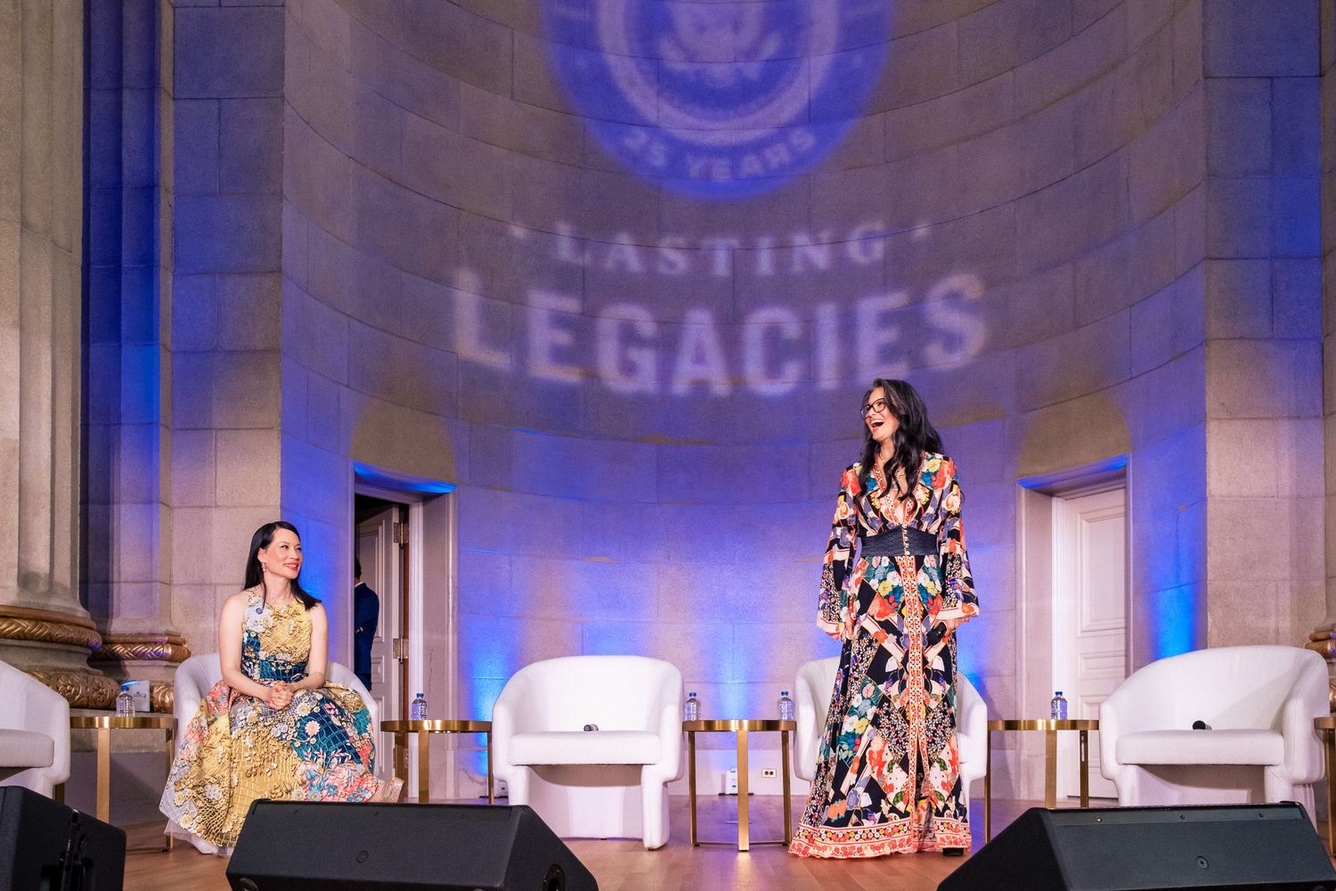 Two women in floral dresses on stage at a Lasting Legacies 25th anniversary event in an elegant venue.