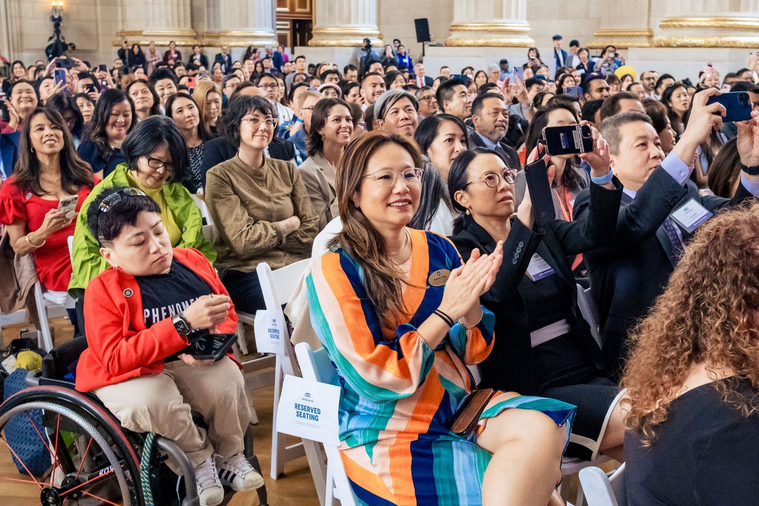 Diverse crowd of attendees applauding at an indoor event, with a woman in colorful sash smiling in the foreground.