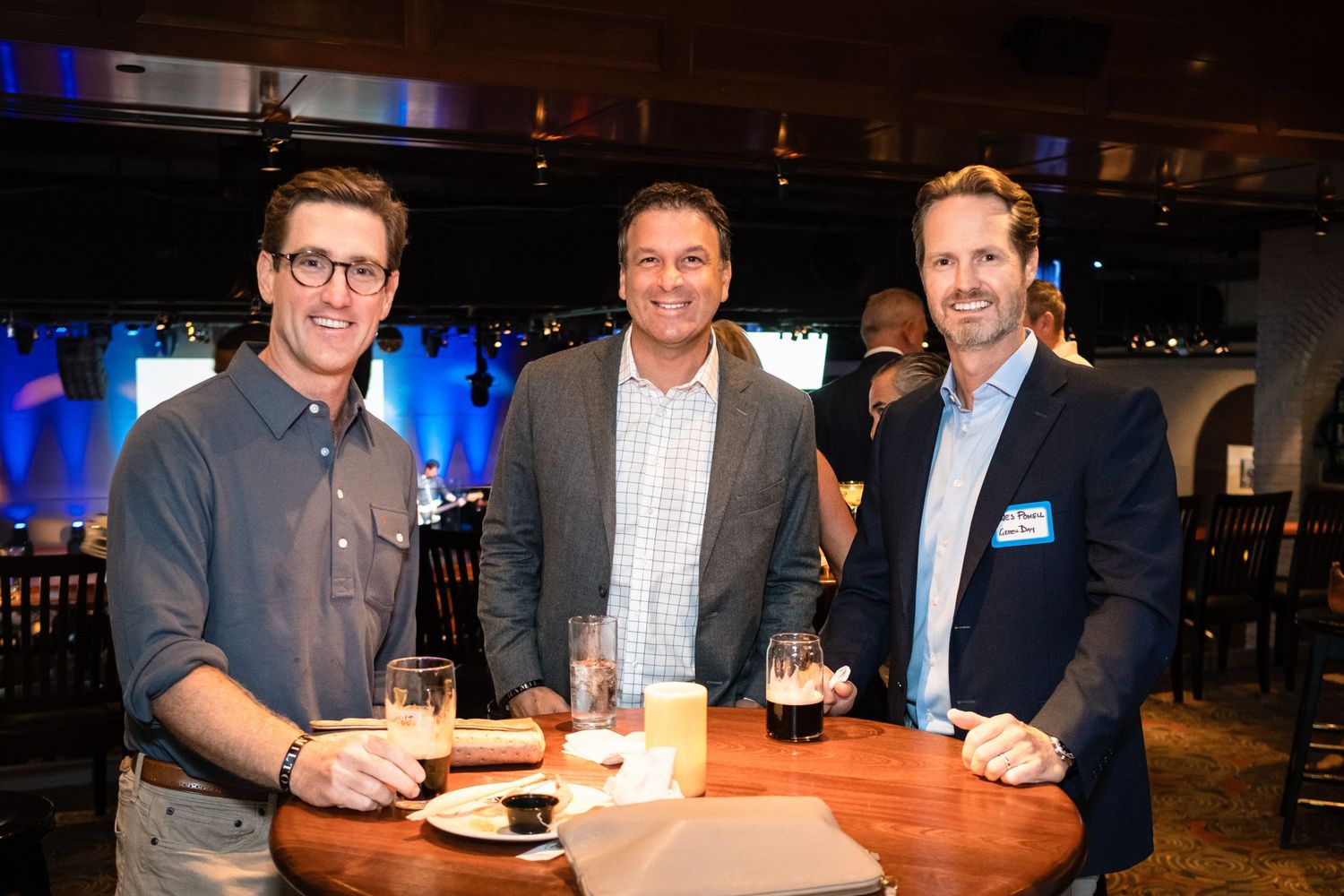 Three smiling men pose together at a high-top table during a corporate networking event in a modern venue.