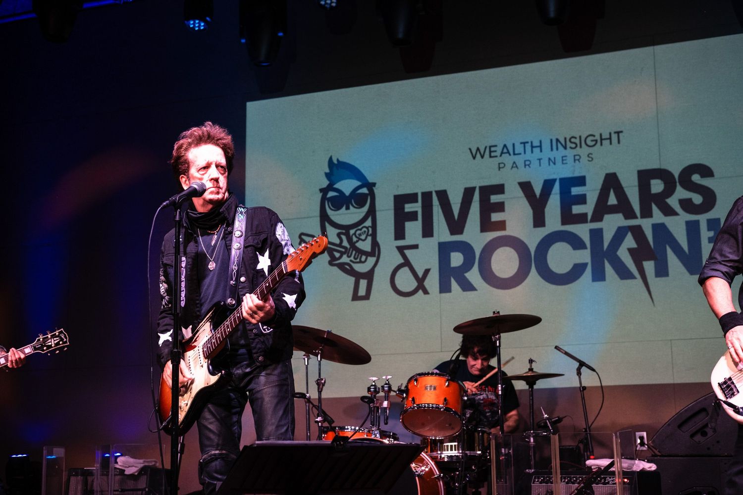 Rock band performs on stage at Wealth Insight Partners Five Years & Rockin anniversary event with guitarist in foreground.