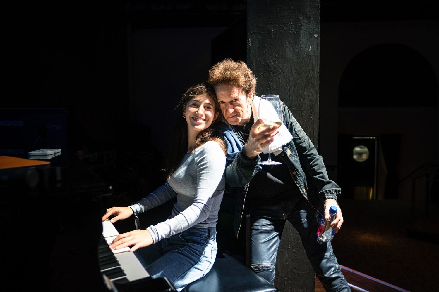 A young woman and man pose together near a piano in a dimly lit performance space.
