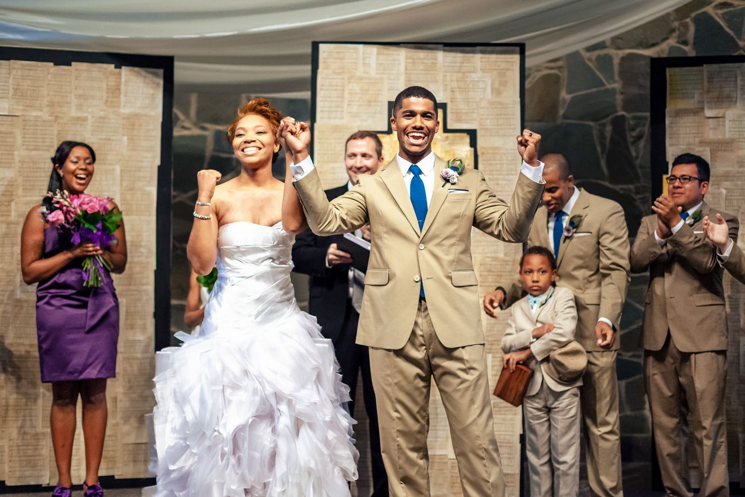 Excited newlywed couple celebrating with raised fists at wedding ceremony exit with bridal party.